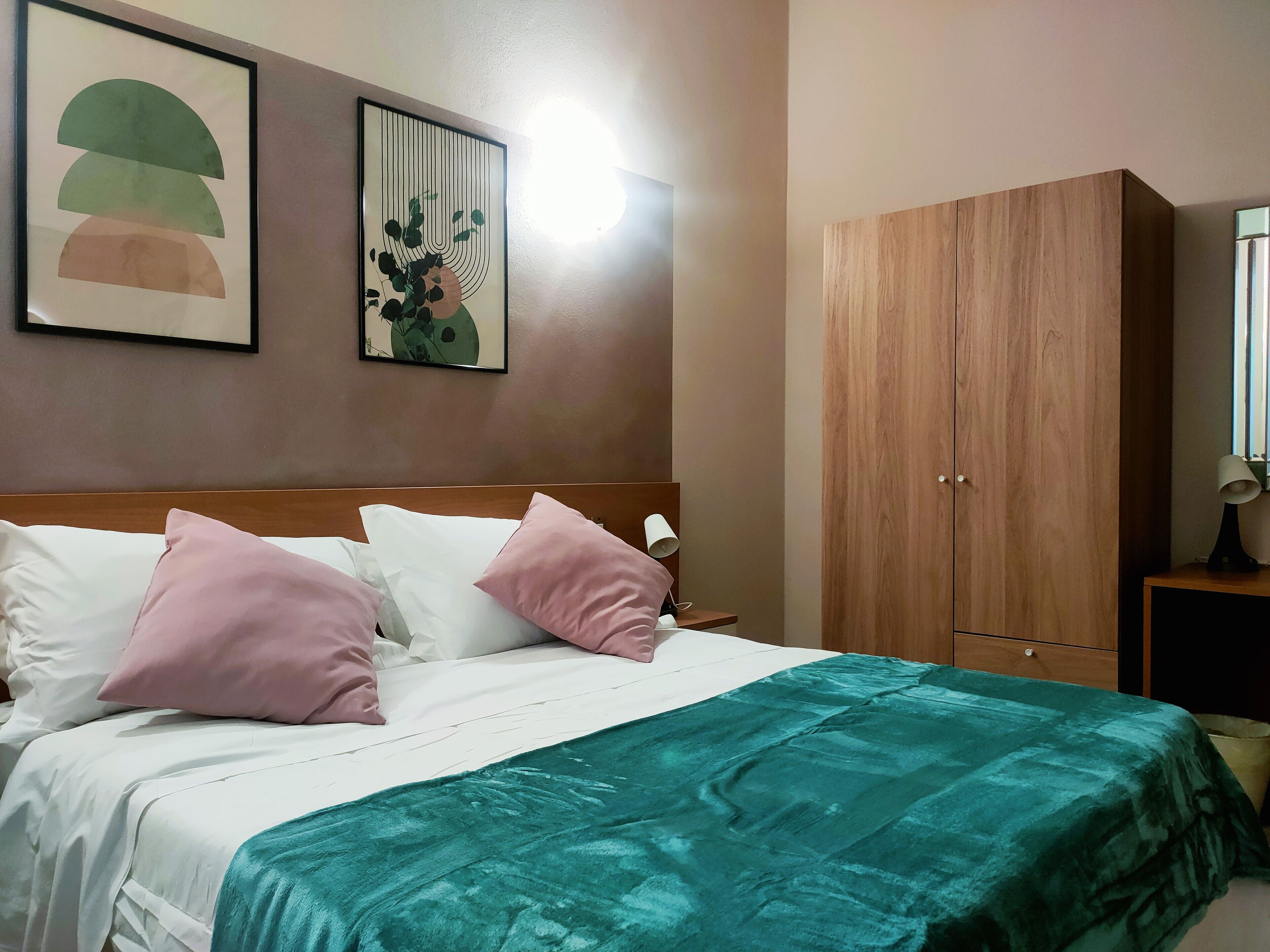 A neatly made bed with white and pink pillows, a teal blanket, and two framed artworks on the wall.