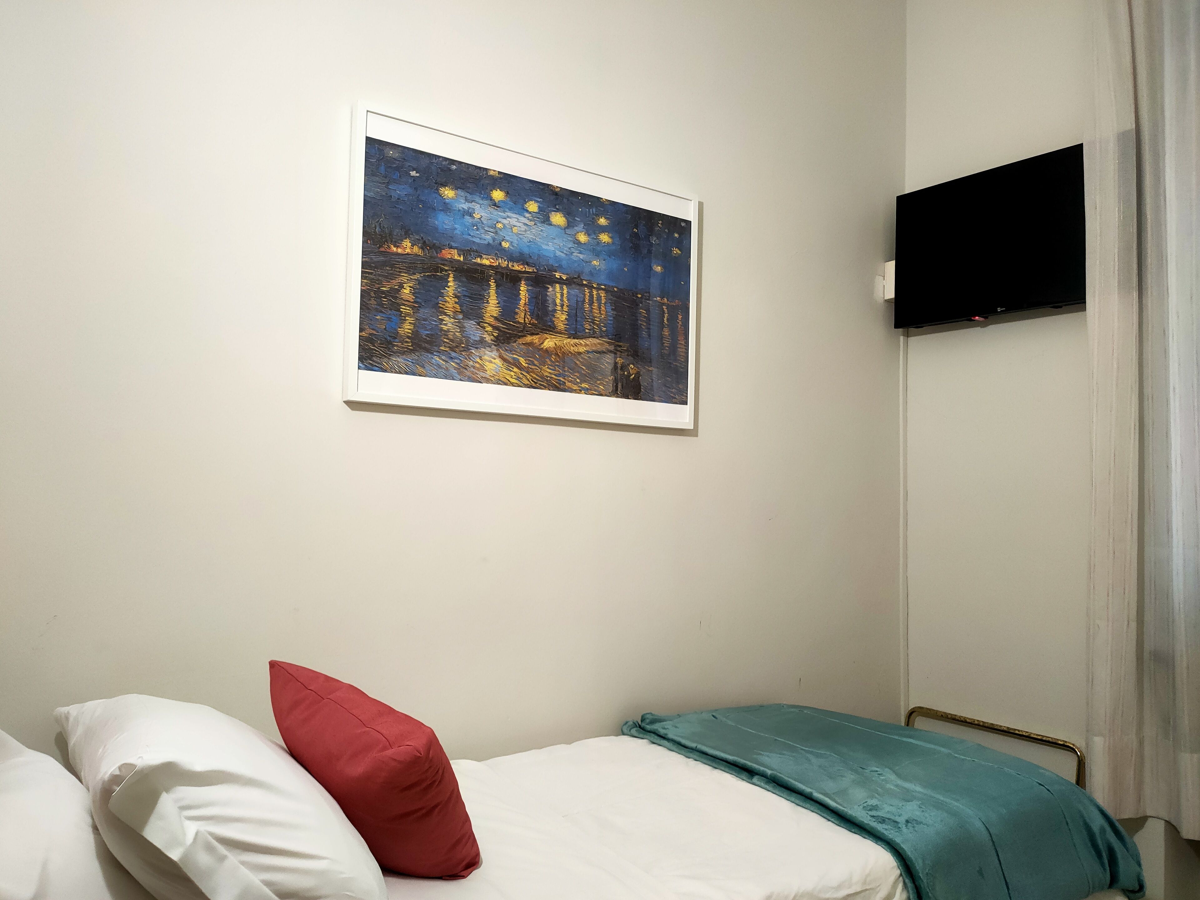 Un dormitorio con cama, almohadas, una colcha verde azulada, un cuadro enmarcado y un televisor en la pared.