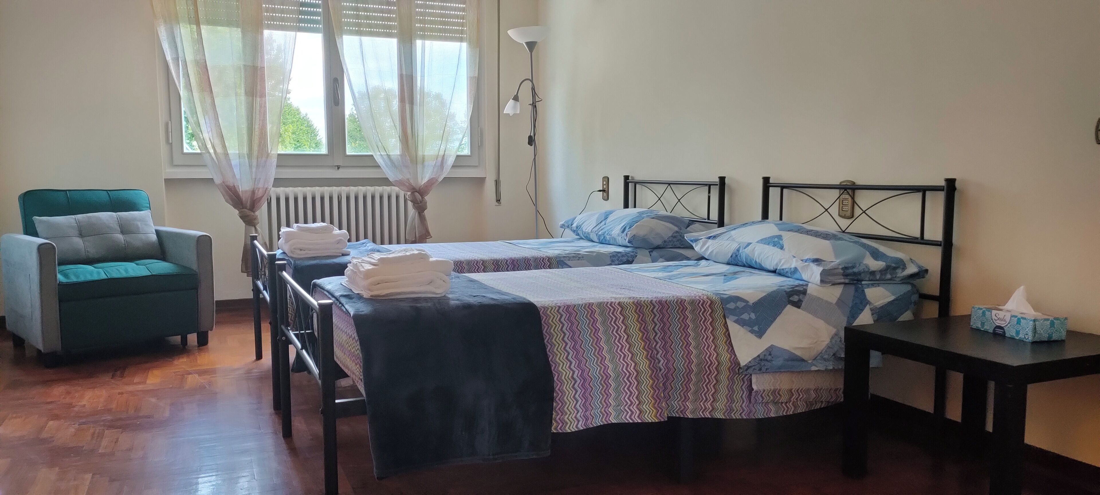 Ein Zimmer mit zwei Betten, einem Sessel, einem Tisch und einem Fenster mit Vorhängen.