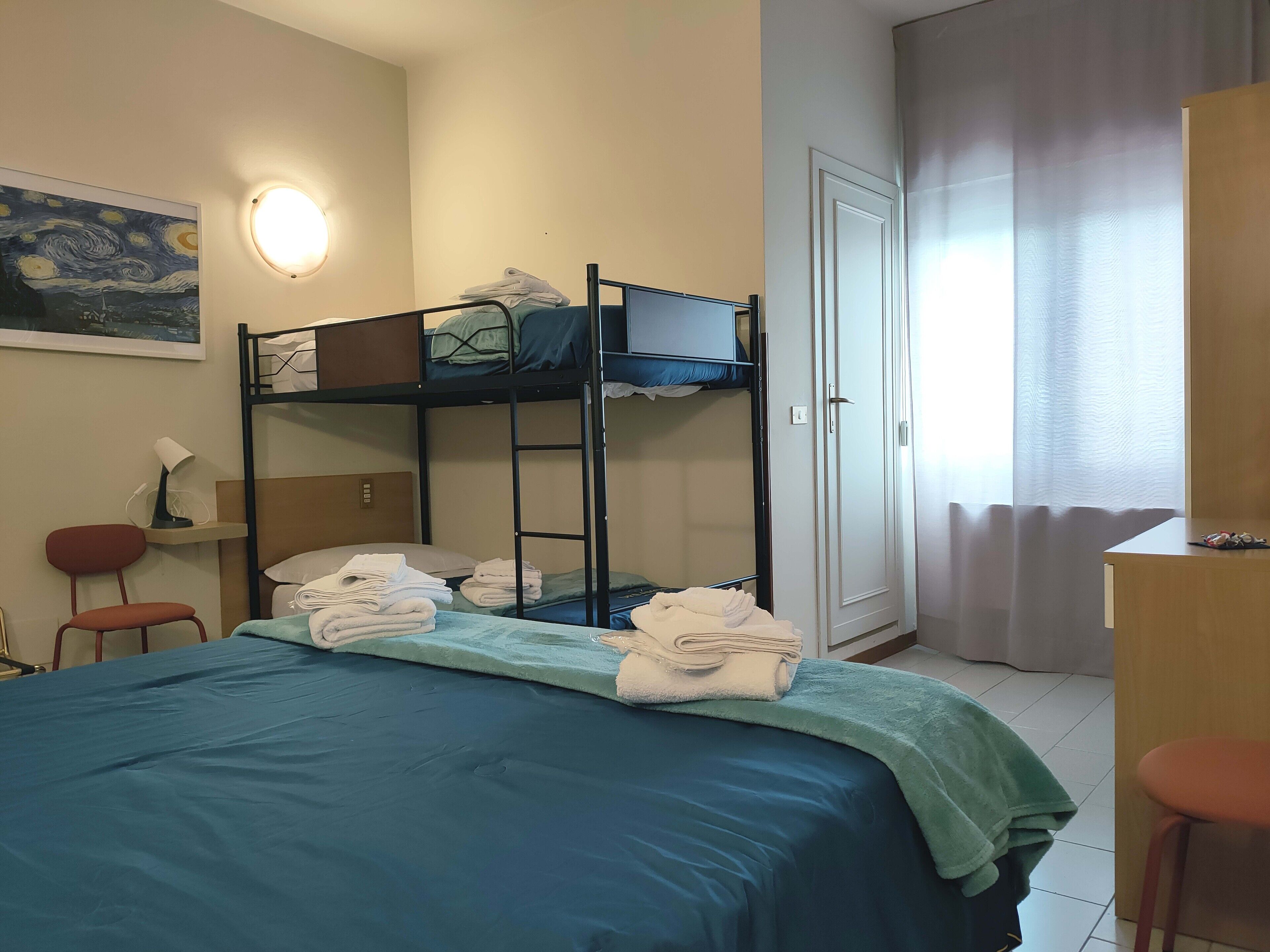 A dormitory room with bunk beds, a desk, a chair, a lamp, and a painting on the wall.