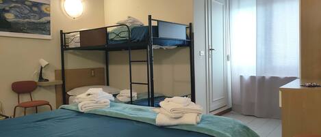 A dormitory room with bunk beds, a desk, a chair, a lamp, and a painting on the wall.