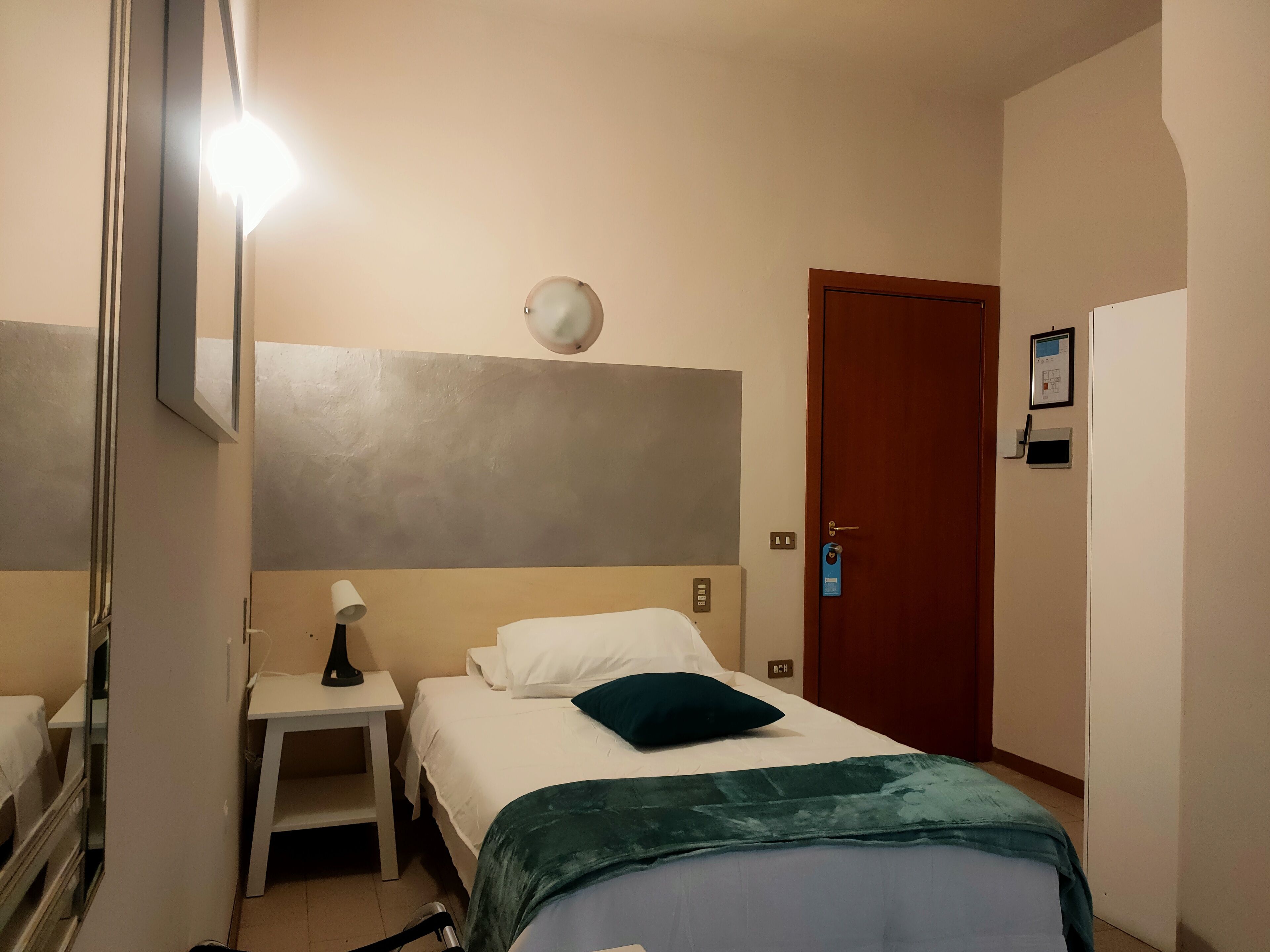 A hotel room with a bed, a bedside table with a lamp, a wall-mounted light, a door with a keycard lock, and a small desk with a chair.