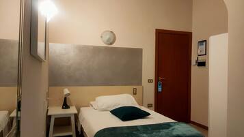 A hotel room with a bed, a bedside table with a lamp, a wall-mounted light, a door with a keycard lock, and a small desk with a chair.