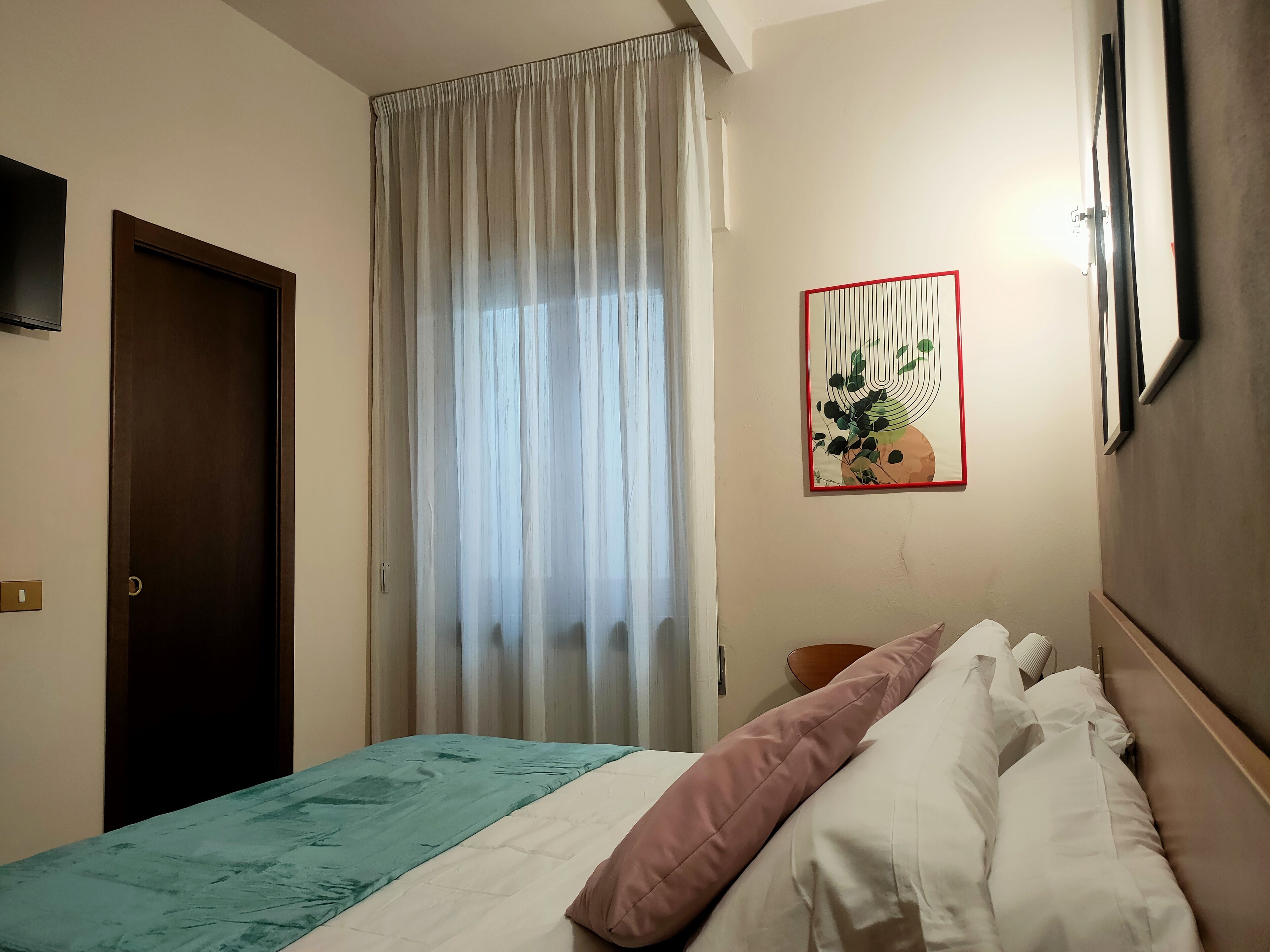 A hotel room with a bed, a window with sheer curtains, a door, and a wall-mounted artwork.