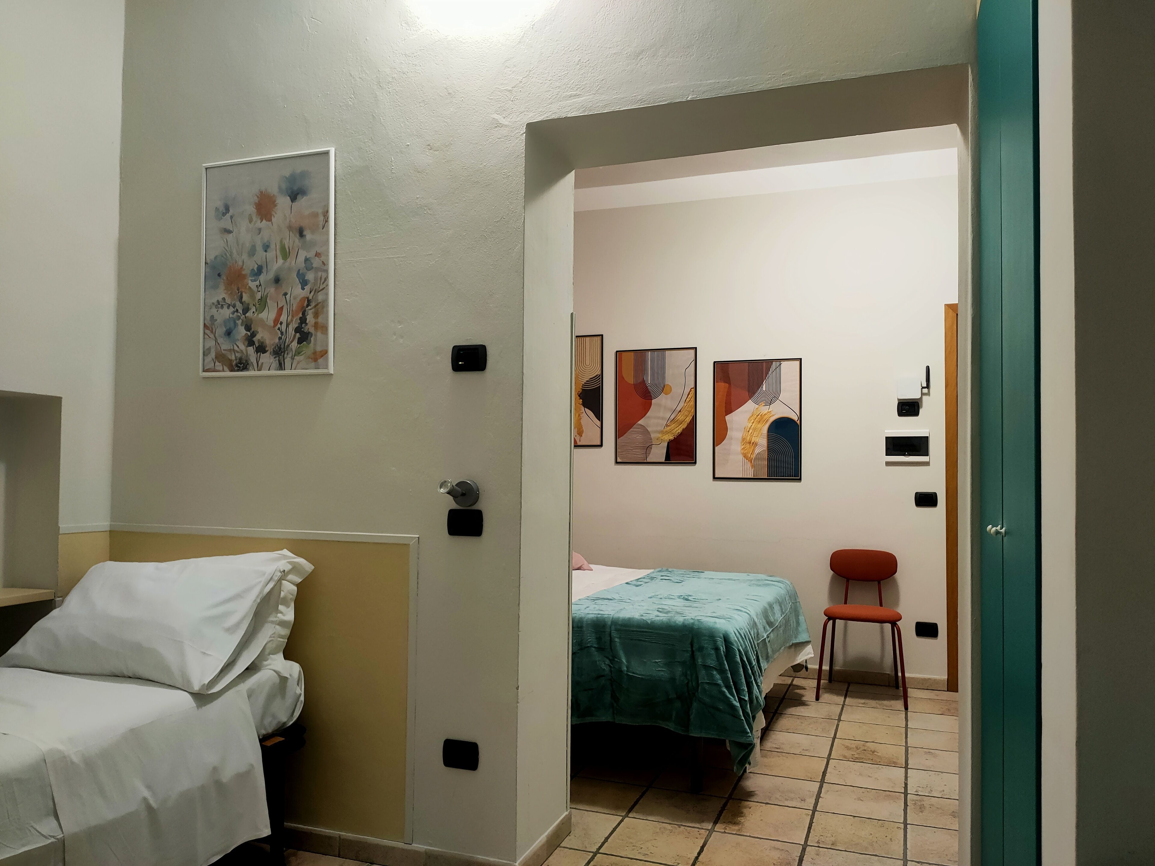 Una camera d'albergo con un letto, una sedia e un tavolino. Tre quadri decorano la parete.