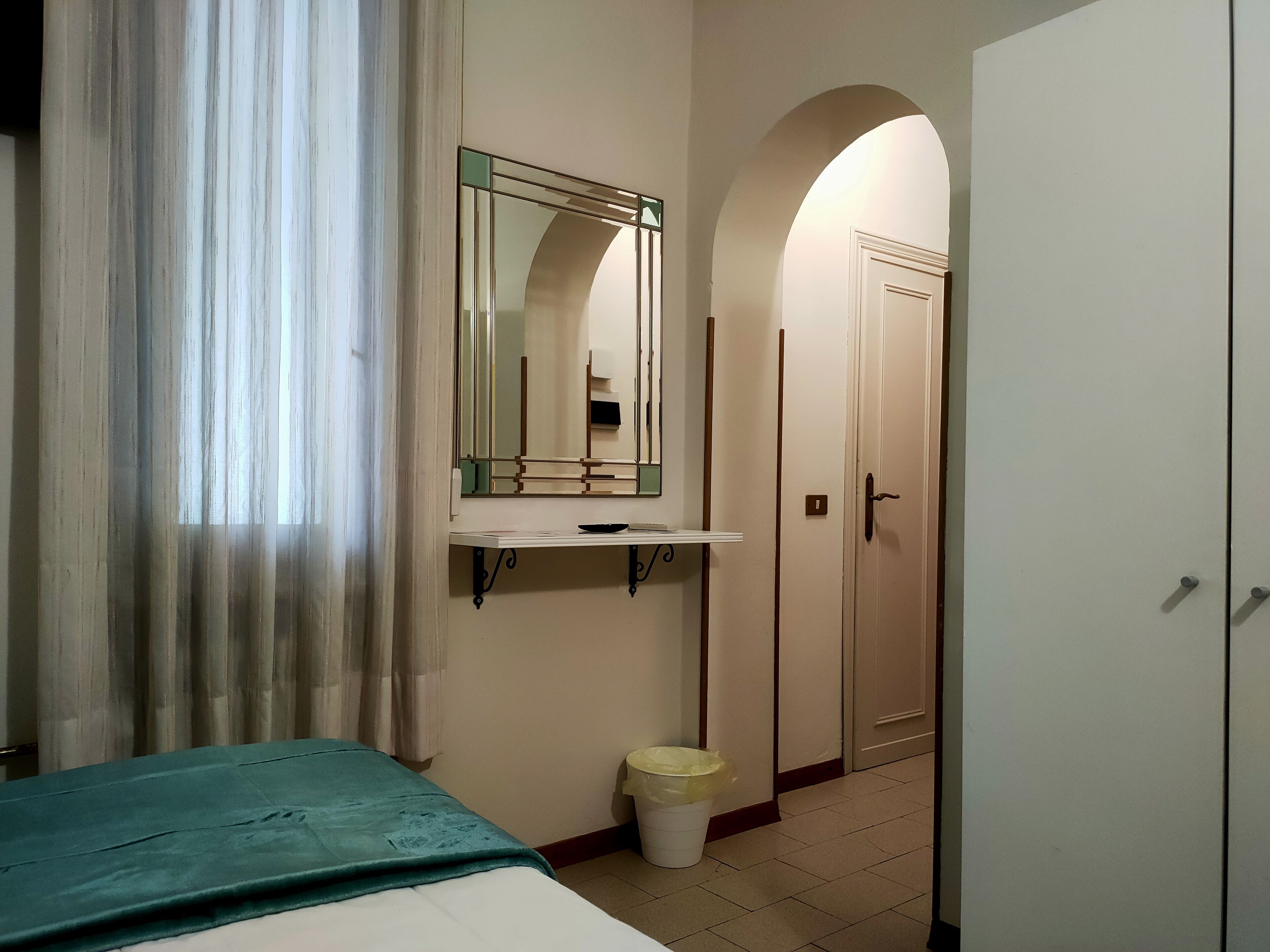 A hotel room with a bed, a bedside table, a mirror, a trash bin, and a wardrobe.