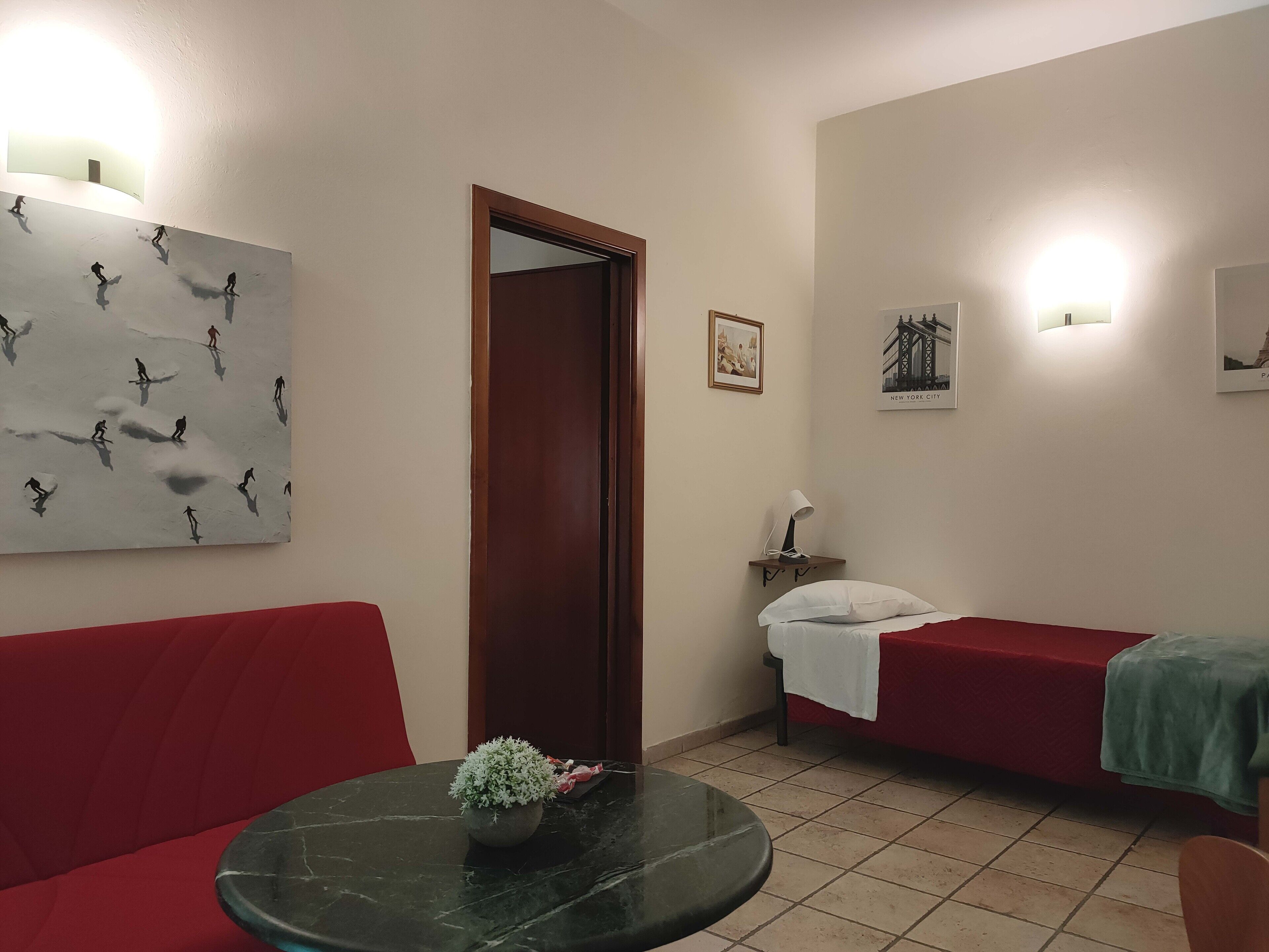 Une pièce avec un canapé rouge, une petite table ronde ornée d’une plante, un lit recouvert d’un couvre-lit vert, et un mur décoré d’un tableau et d’une photographie encadrée.