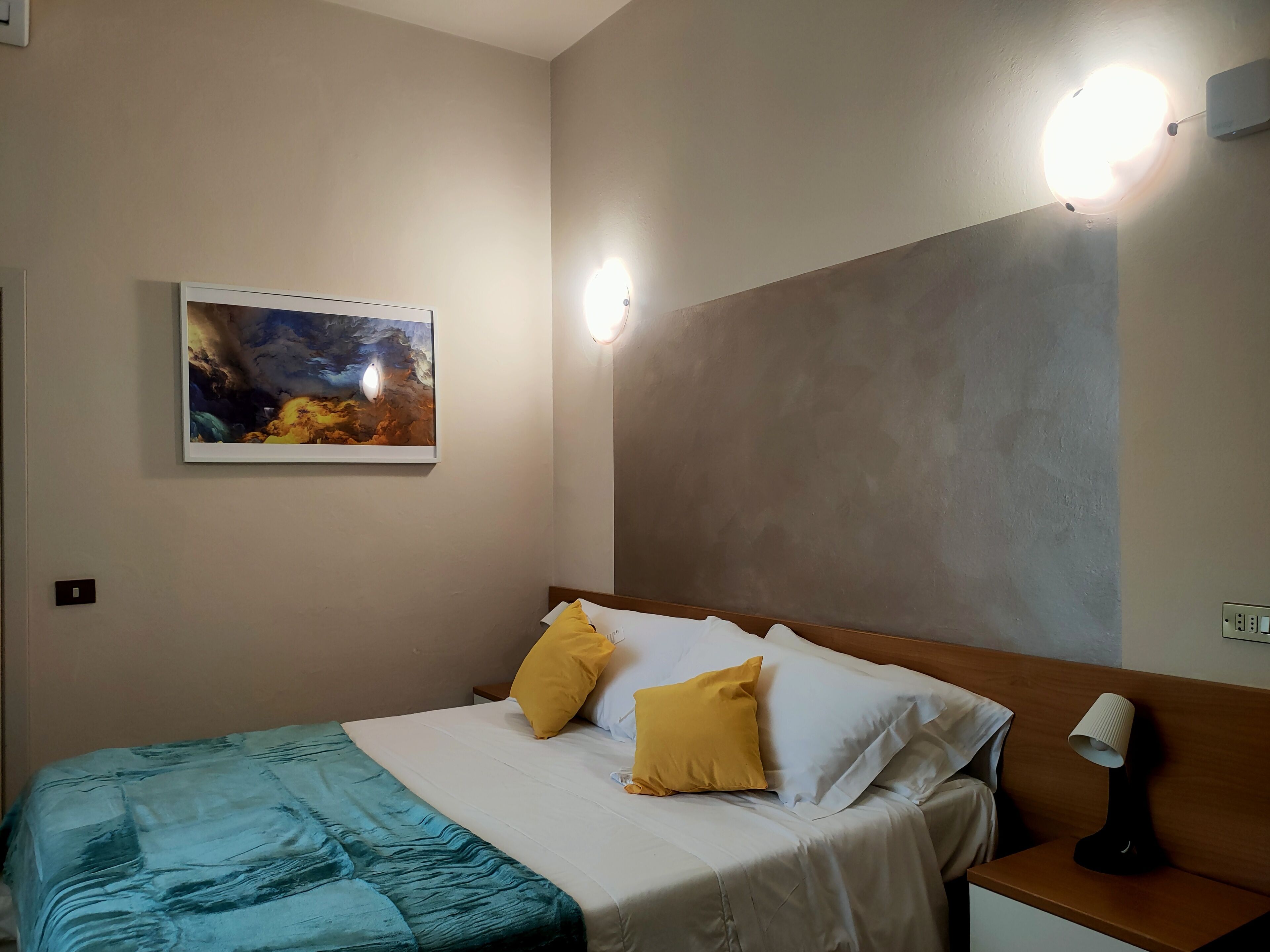 Une chambre à coucher comprenant un lit, deux lampes de chevet, une table de chevet, un luminaire fixé au mur et un tableau encadré.