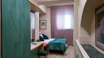 Camera d'albergo compatta con un letto, una scrivania e una sedia. Si trova un armadio su un lato e una finestra con tende.
