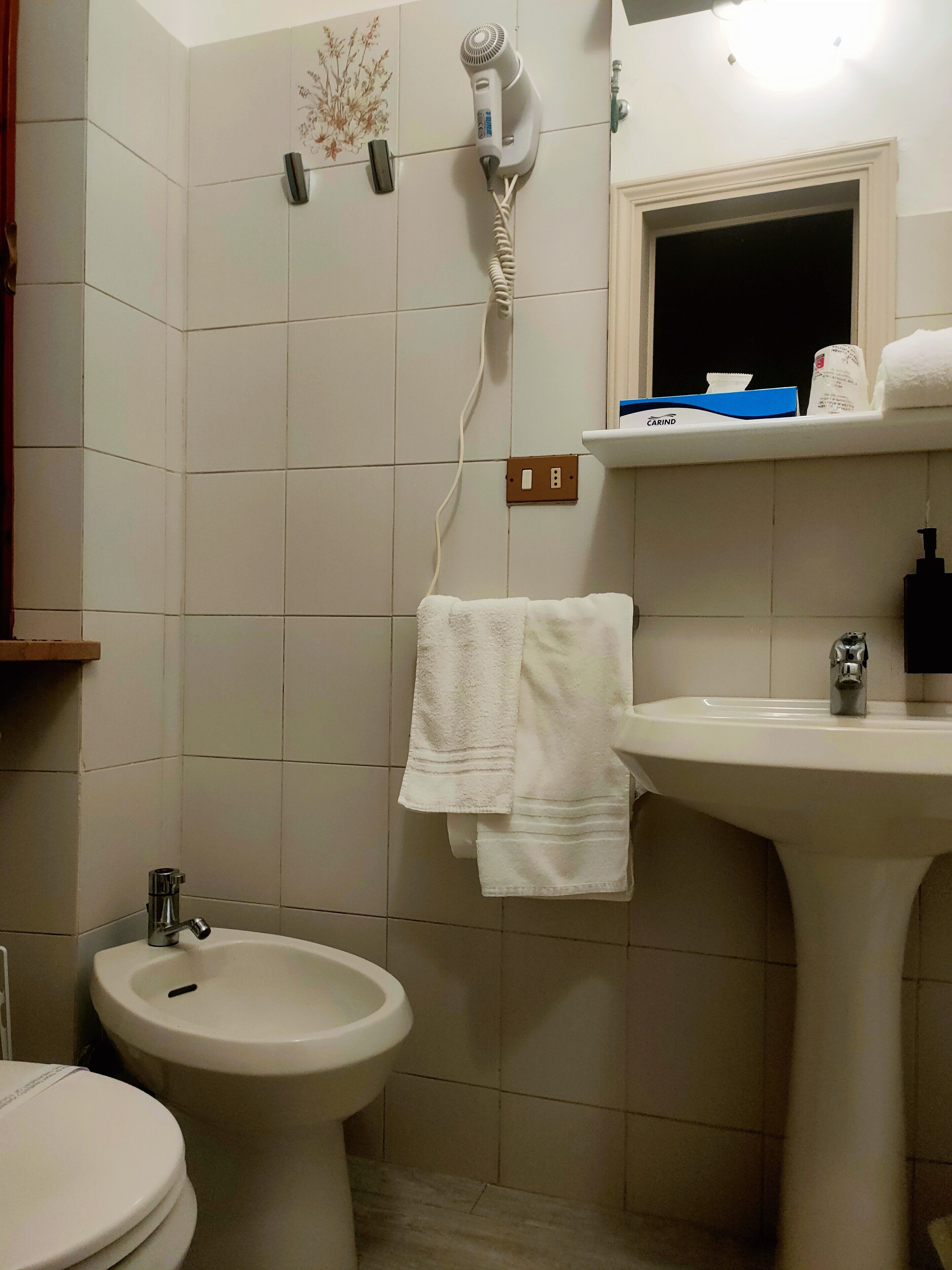 A bathroom with a toilet, bidet, sink, and a hairdryer mounted on the wall.
