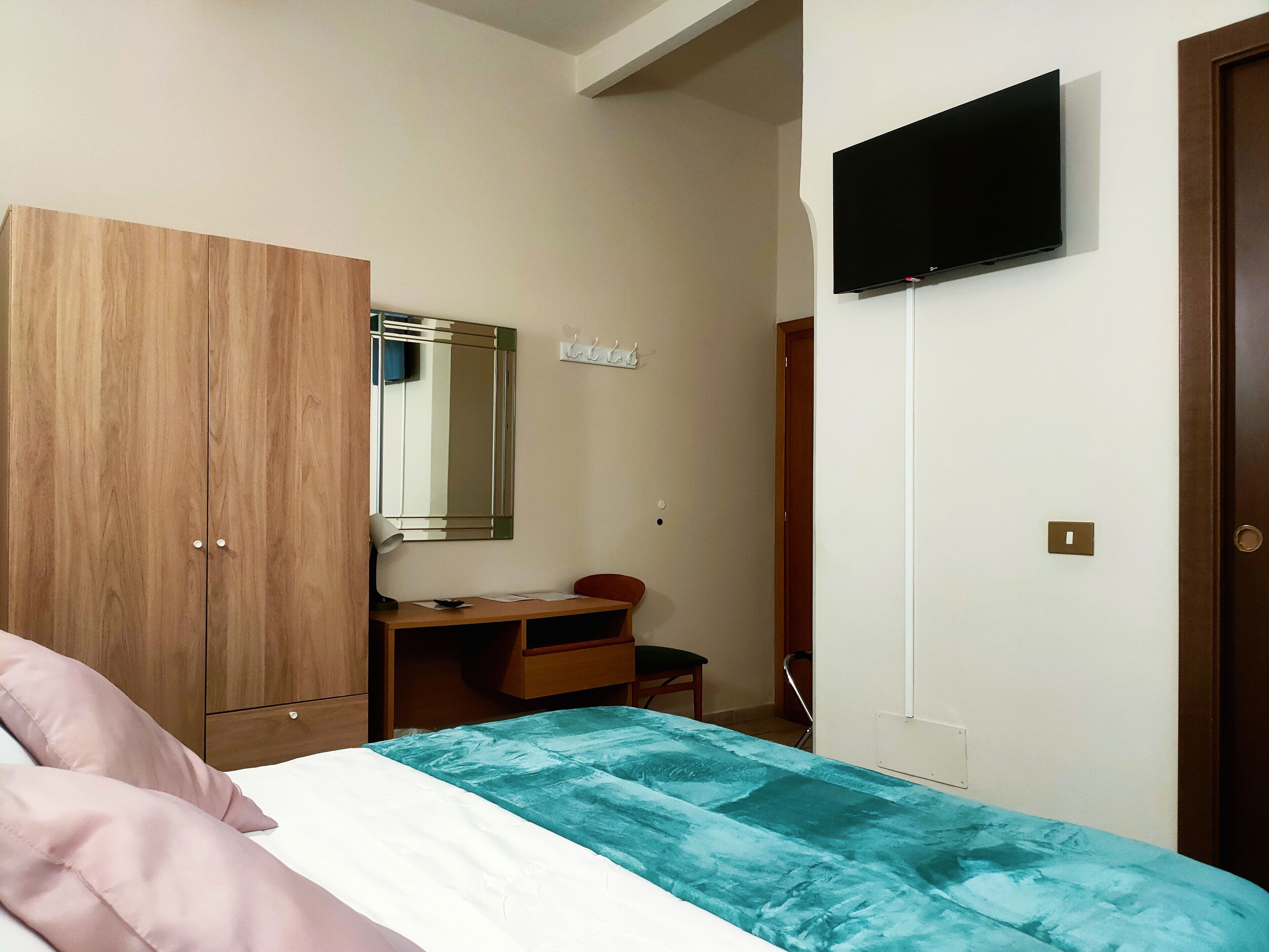 Un dormitorio con cama, un armario de madera, un escritorio con silla, televisor montado en la pared y una puerta.