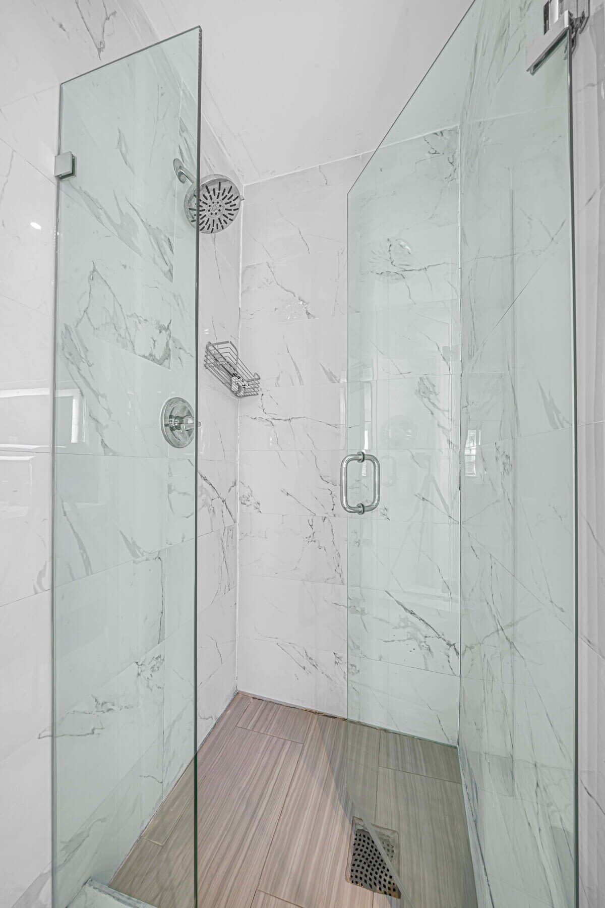 A glass-enclosed shower with marble walls and a wooden floor.