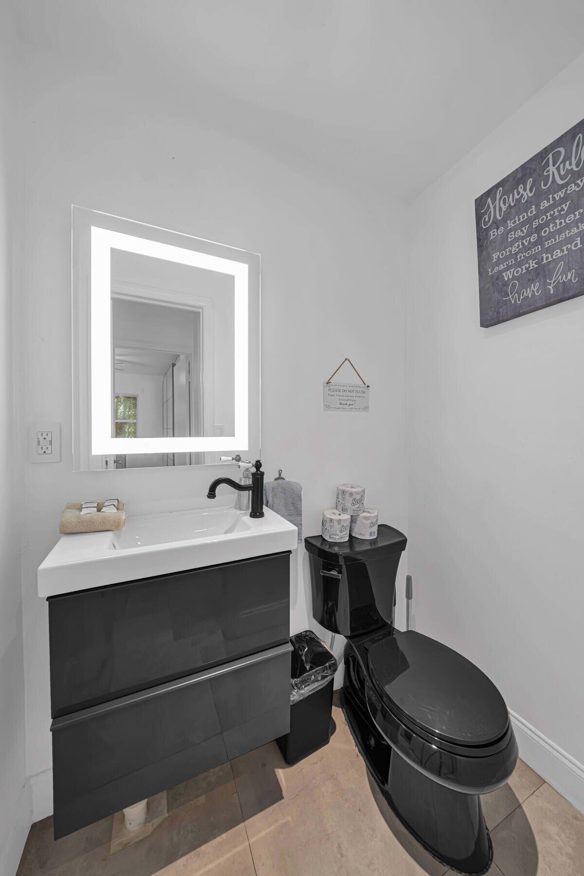 Ein modernes Badezimmer mit einem weißen Waschtisch, einem schwarzen Waschbecken und einer Toilette. Über dem Waschbecken hängt ein Spiegel und an der Wand befindet sich ein Schild mit motivierendem Text.