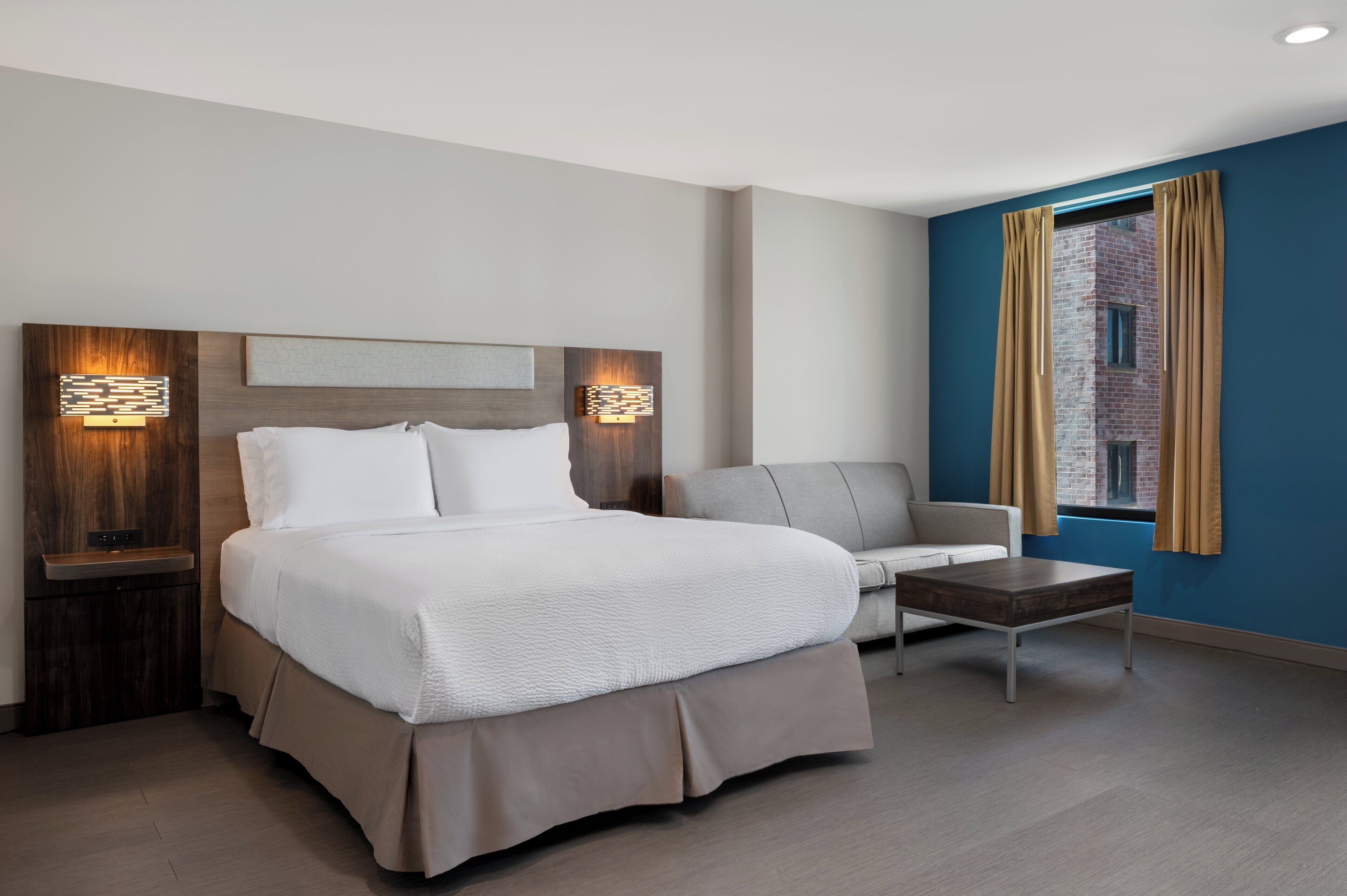 Una habitación de hotel moderna con una cama grande, un sofá, una mesita y una ventana con cortinas.
