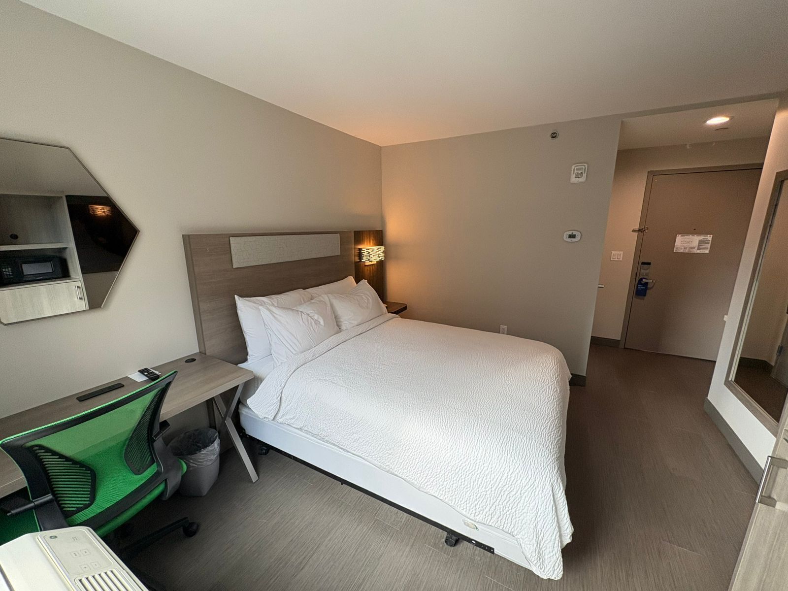 A hotel room with a bed, a desk with a chair, a trash bin, and a wall-mounted microwave.