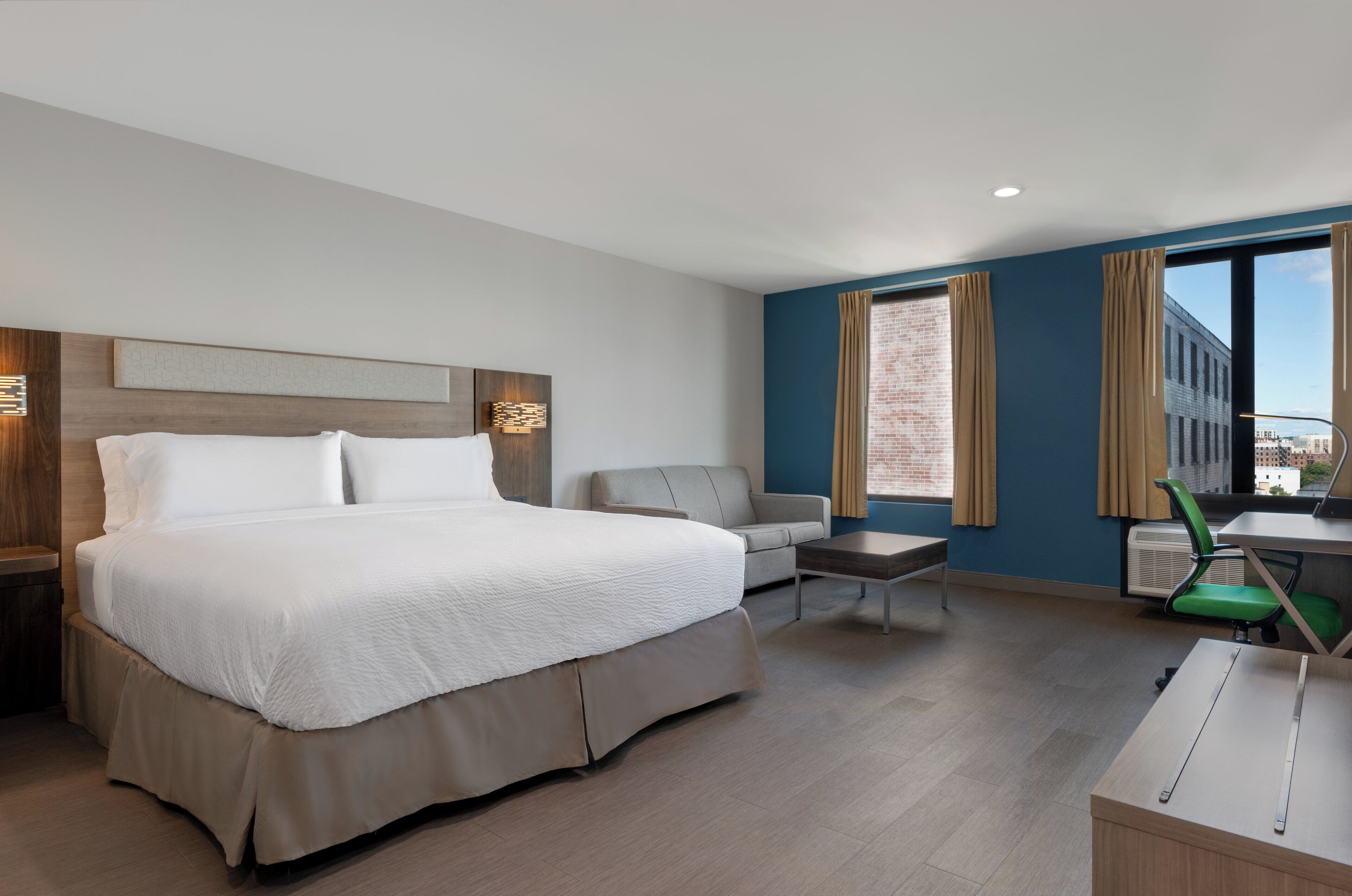 A modern hotel room with a large bed, a desk, a chair, and a view of the city through large windows.