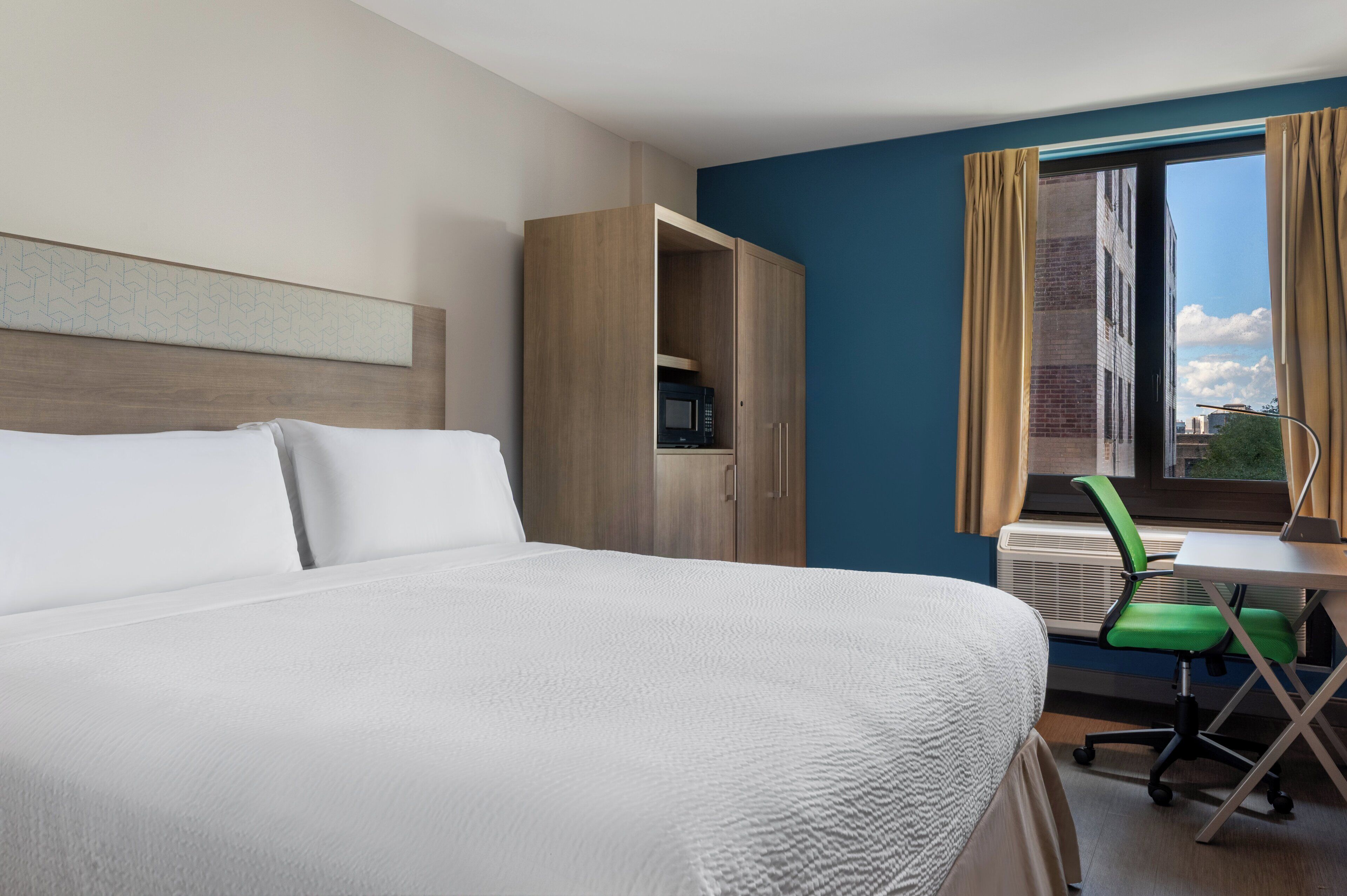 A hotel room with a bed, a desk with a chair, an air conditioner, and a view of the city through a window.