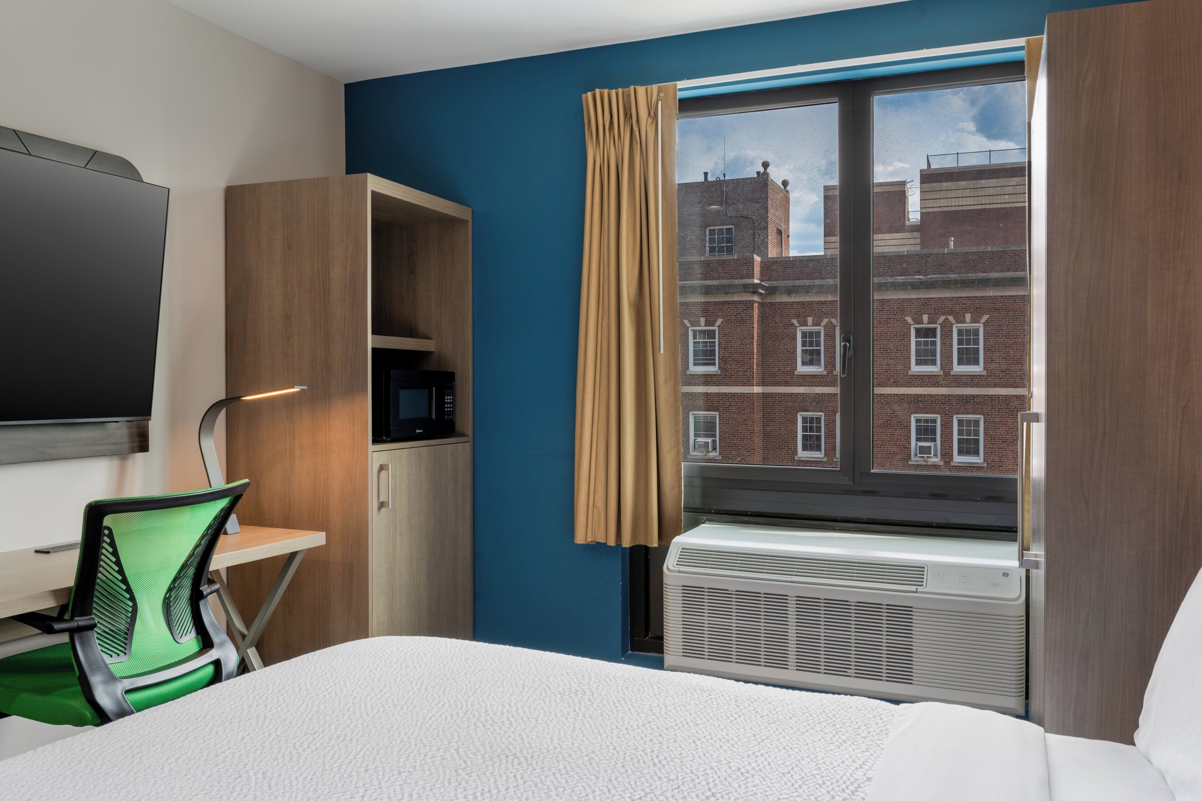 A hotel room with a bed, a desk with a computer, a TV, a window with curtains, and a view of a brick building.