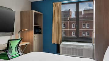 A hotel room with a bed, a desk with a computer, a TV, a window with curtains, and a view of a brick building.