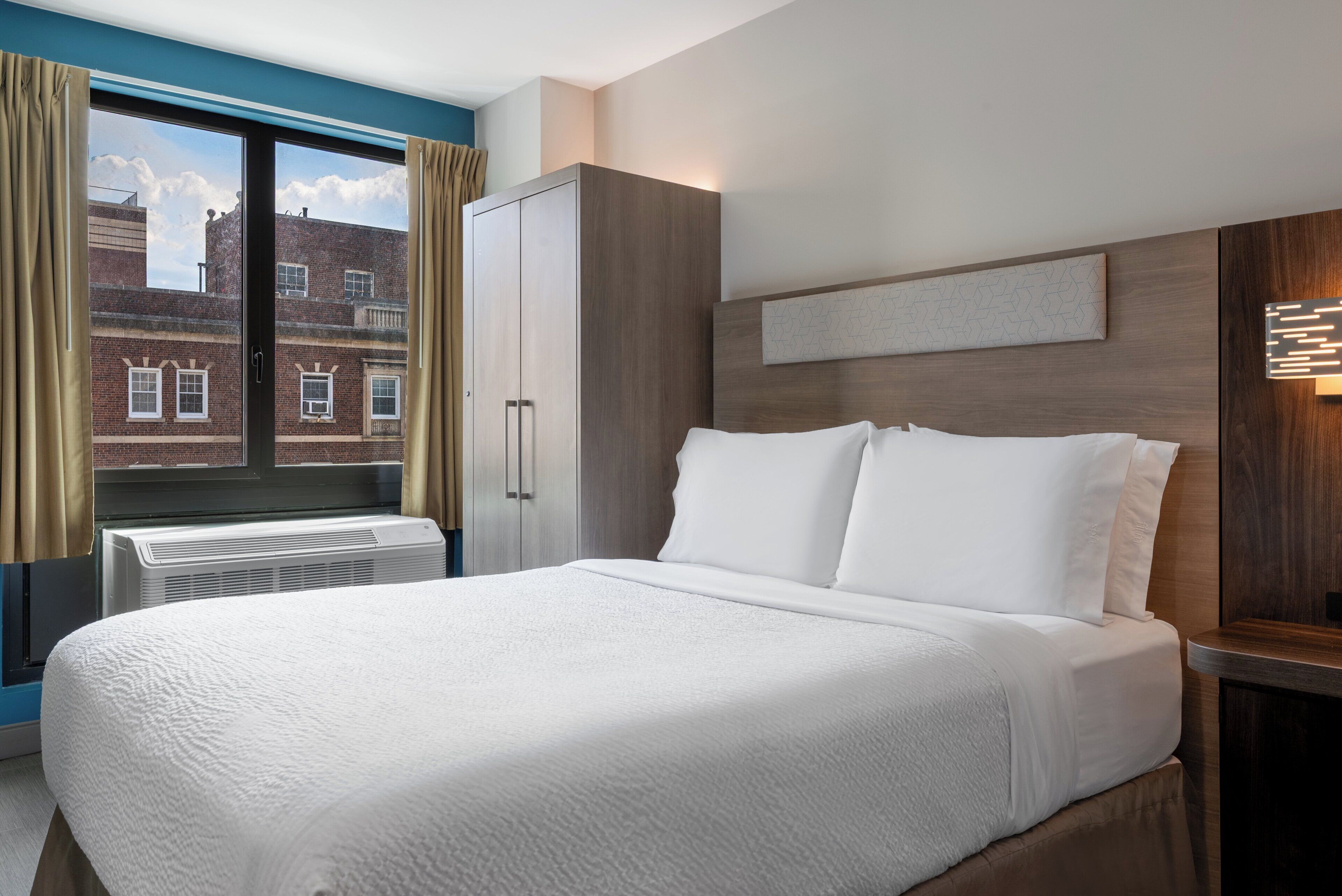 Ein Hotelzimmer mit einem großen Bett, einem Kleiderschrank, einem Fenster mit Vorhängen und Blick auf Gebäude.