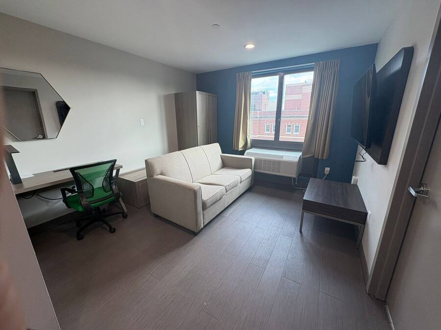 Een kamer met een bank, een bureau met stoel, een aan de muur bevestigde televisie en een raam met gordijnen.