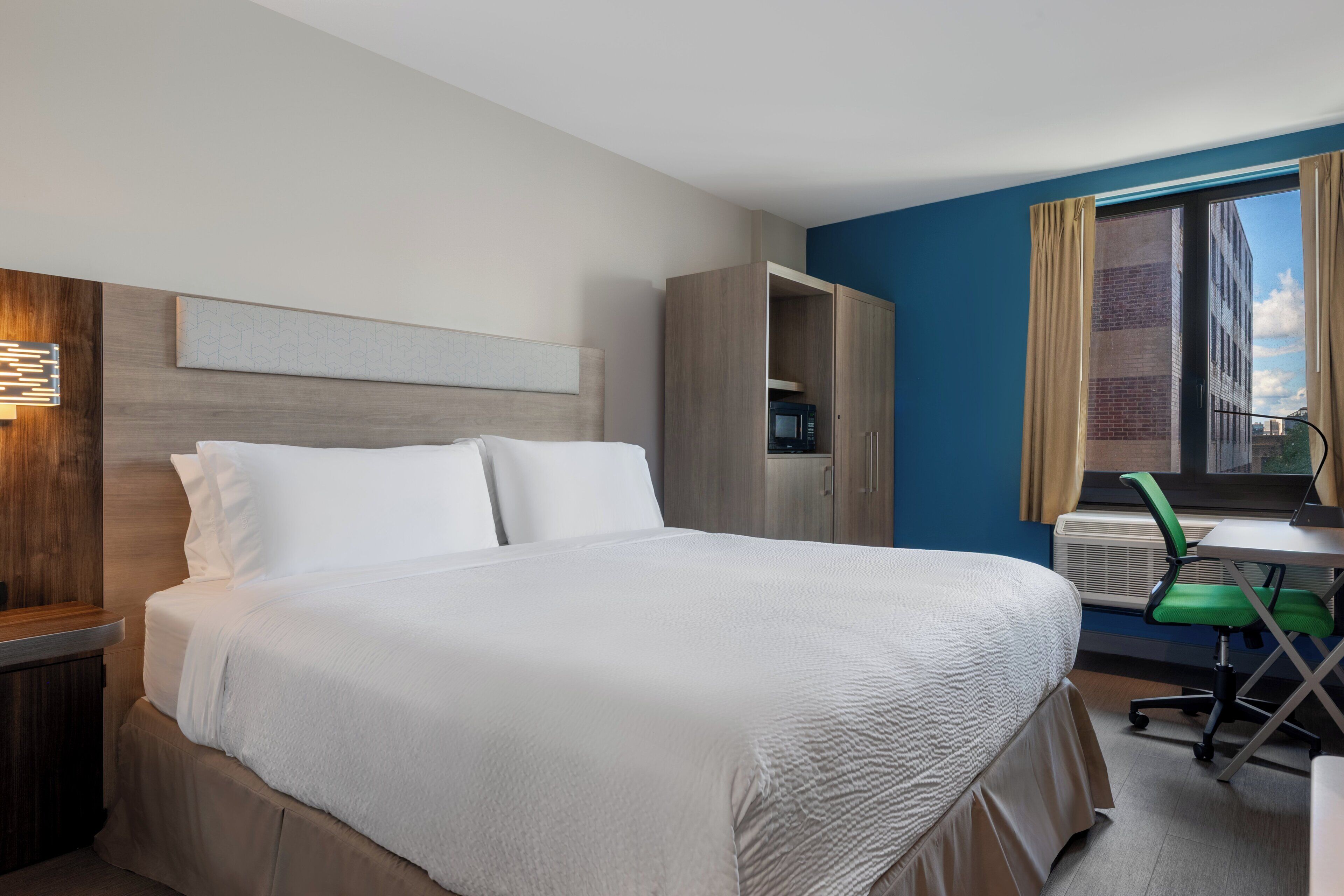 A hotel room with a large bed, a desk with a chair, a wardrobe, and a window with curtains.