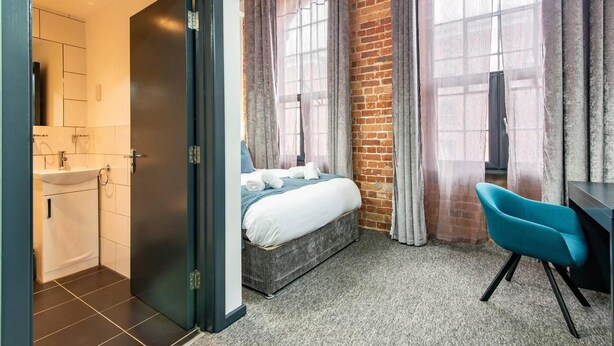A bedroom with a bed, a desk, a chair, and a bathroom with a sink and mirror. - Stylish 1-bed Studio - Heart of Nottingham (Nottingham)