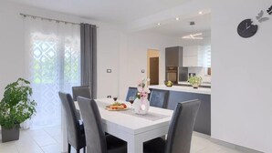 A modern dining area with a white table, black chairs, and a vase with flowers. A kitchen is visible in the background with white cabinets and a clock on the wall. - Lovely home in Svetvincenat (Svetvincenat)