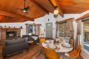 Una sala de estar acogedora con chimenea, techo de madera y un comedor con sillas amarillas y una mesa preparada para cuatro.