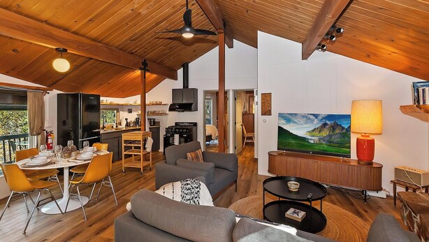 Een ruime woonkamer met een hoge plafond, houten balken, een eettafel met stoelen, een bank, een televisie en een schilderij aan de muur.
