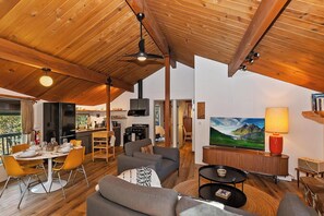 A spacious living area with a high ceiling, wooden beams, a dining table with chairs, a sofa, a TV, and a painting on the wall.