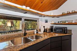 Una cocina con encimeras de granito, pileta de acero inoxidable y microondas. Hay repisas de madera sobre la encimera y una ventana con vista a árboles.