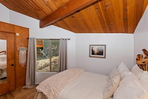Un dormitorio con techo de madera, cama con ropa de cama blanca, un atrapasueños, un cuadro enmarcado y vistas al exterior.