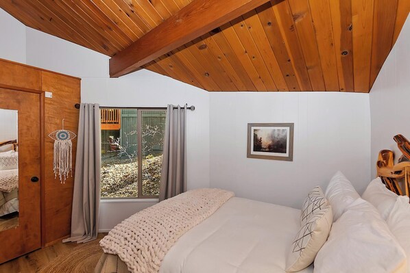 A bedroom with a wooden ceiling, a bed with white bedding, a window with curtains, and a door with a wooden frame.
