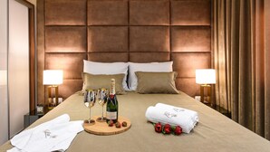 A hotel room with a tufted headboard, two bedside lamps, a bed with white and beige bedding, a wooden tray with champagne glasses and a bottle, and rolled white towels with a monogram.