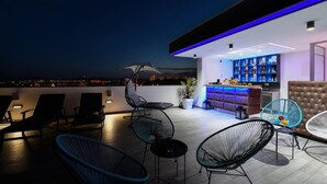 A rooftop bar with a bar counter, seating area with chairs, and a view of the city at night.