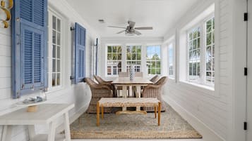 A room with a white wooden interior, a ceiling fan, a table with wicker chairs, and large windows.