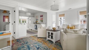 Een ruime woonkamer met een witte bank, een bijzettafeltje en een kleurrijk vloerkleed. Achtergrond is een keuken met witte kasten en een eettafel.