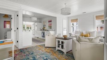 A spacious living room with a white sofa, a coffee table, and a colorful rug. There is a kitchen area in the background with white cabinets and a dining table.