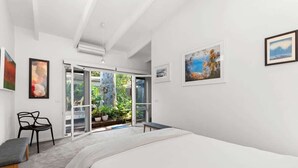 A bedroom with a bed, a chair, a desk, and a sliding glass door leading to a balcony with plants.