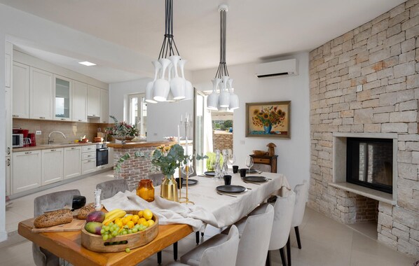 A modern kitchen with white cabinets, a brick accent wall, a dining area with a white table set, and a fireplace. - Villa Bincila Labinci (Porec)