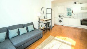 A modern apartment with a grey sofa, a small dining area, and a kitchen with white cabinets and a washing machine. - Charming 2-bed Apartment in London (London)