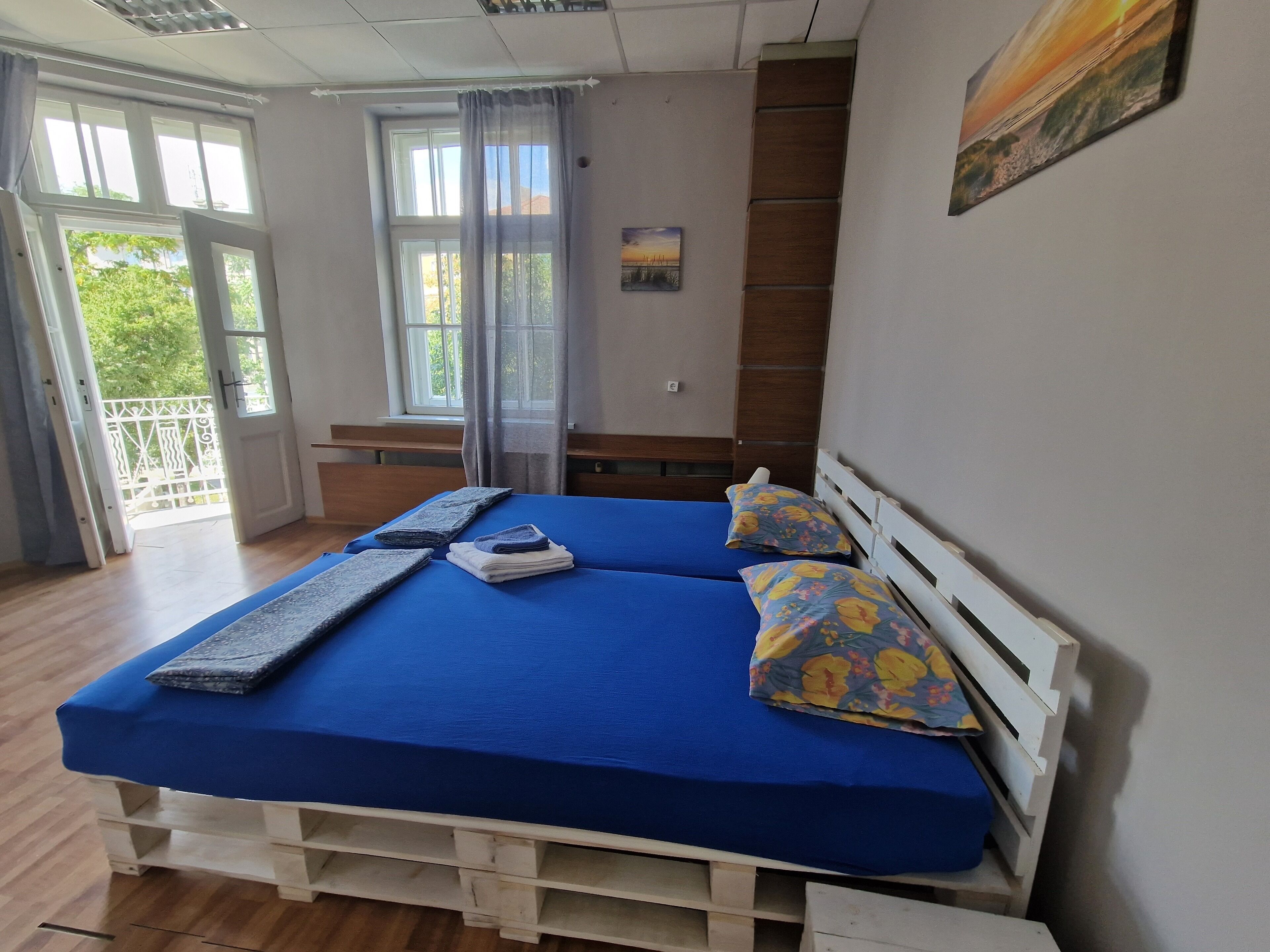 A double bed with blue bedding and floral pillows, a wooden headboard, and a view of a balcony through a window with curtains.