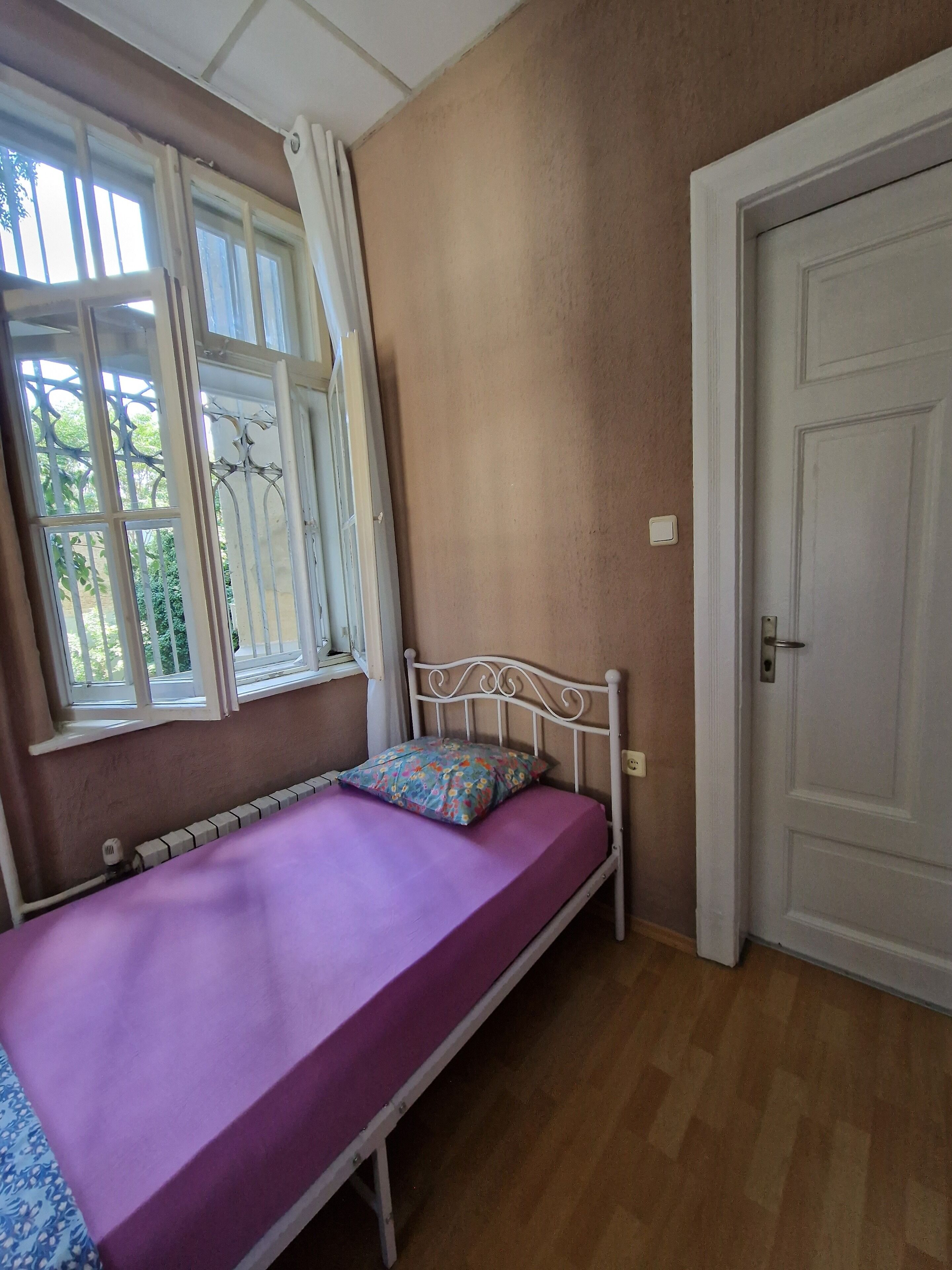 Una cama individual con colchón morado, estructura metálica blanca y almohada estampada. Se ve una ventana con cortinas blancas y un radiador debajo. La habitación tiene paredes color beige y suelo de madera.