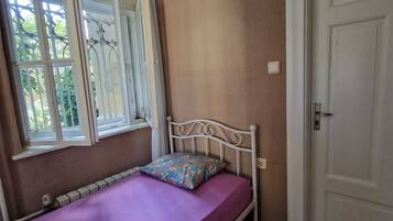 Una cama individual con colchón morado, estructura metálica blanca y almohada estampada. Se ve una ventana con cortinas blancas y un radiador debajo. La habitación tiene paredes color beige y suelo de madera.