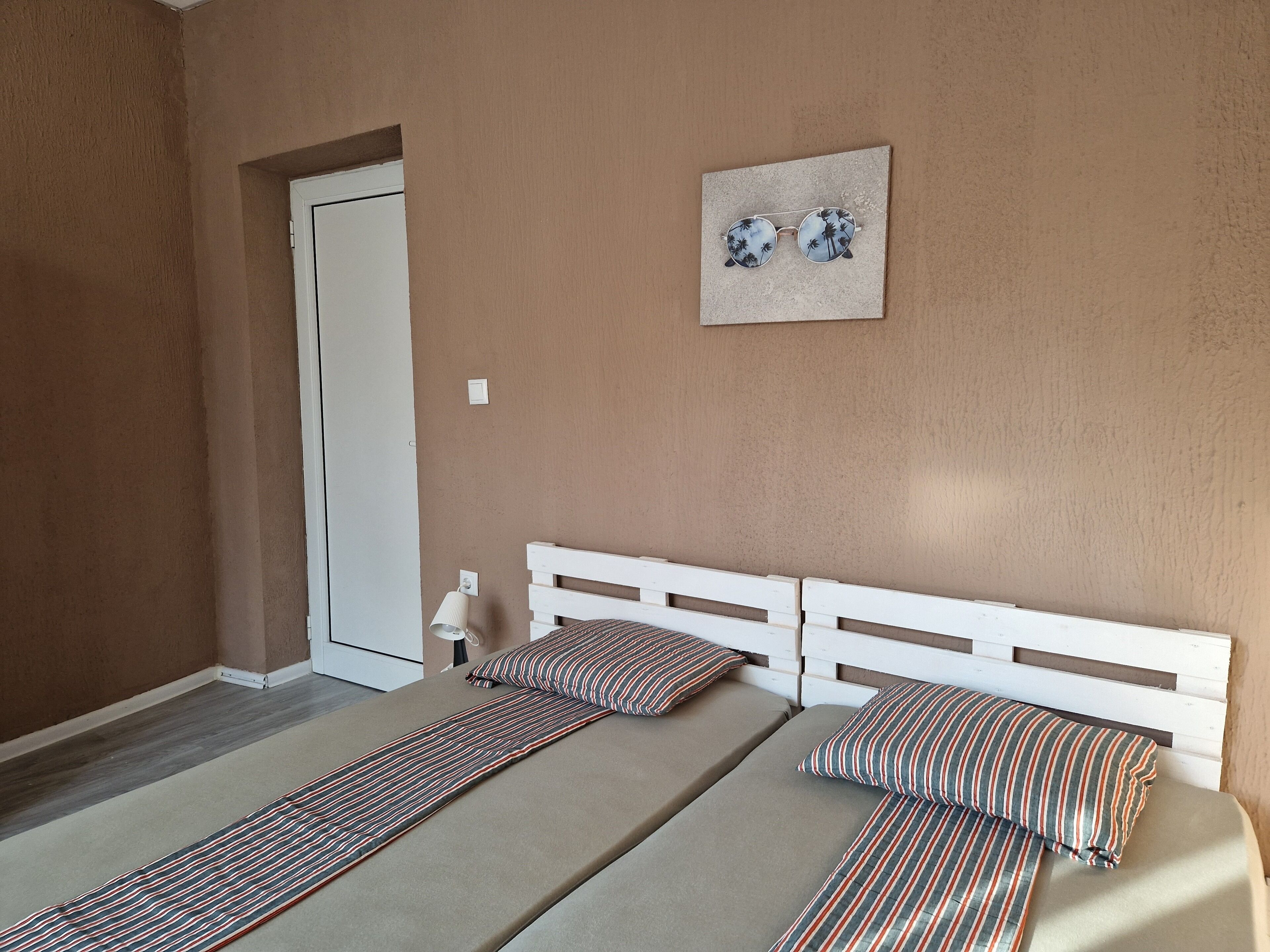 A room with two beds, a white headboard, a door, a wall-mounted light, and a framed picture.