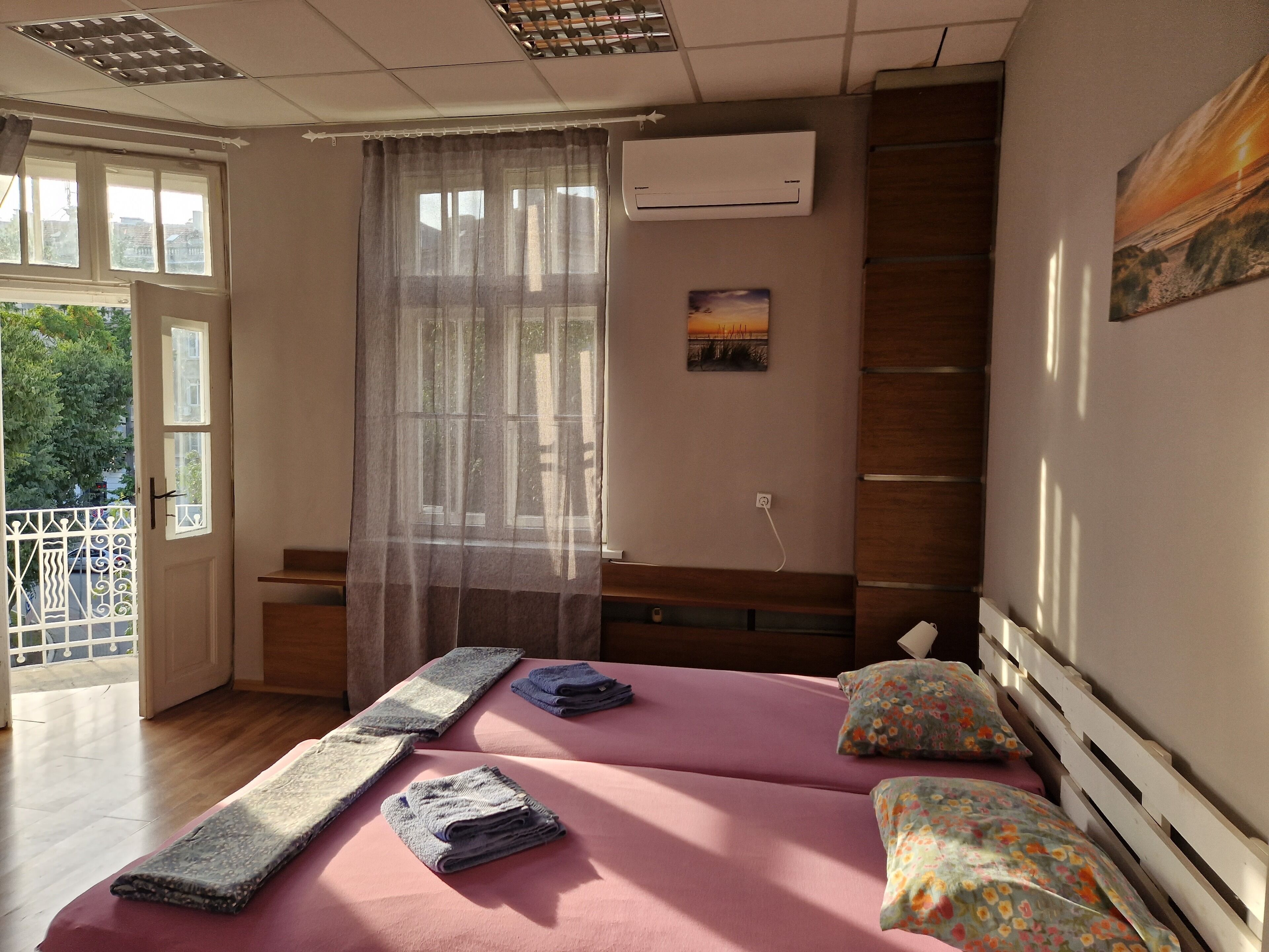 Un dormitorio con dos camas, una ventana con cortinas traslúcidas, un equipo de aire acondicionado y un cuadro en la pared.