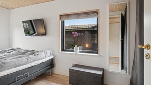 Een slaapkamer met een bed, een klein nachtkastje, een raam met uitzicht op een houten gebouw, een spiegel en een aan de muur bevestigde televisie.