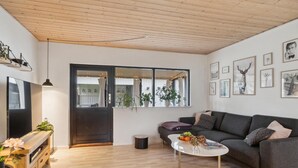 Een moderne woonkamer met een donkergrijze bank, een witte salontafel en een tv op een houten dressoir. Er staan verschillende potplanten en er hangen kunstwerkjes aan de muren.