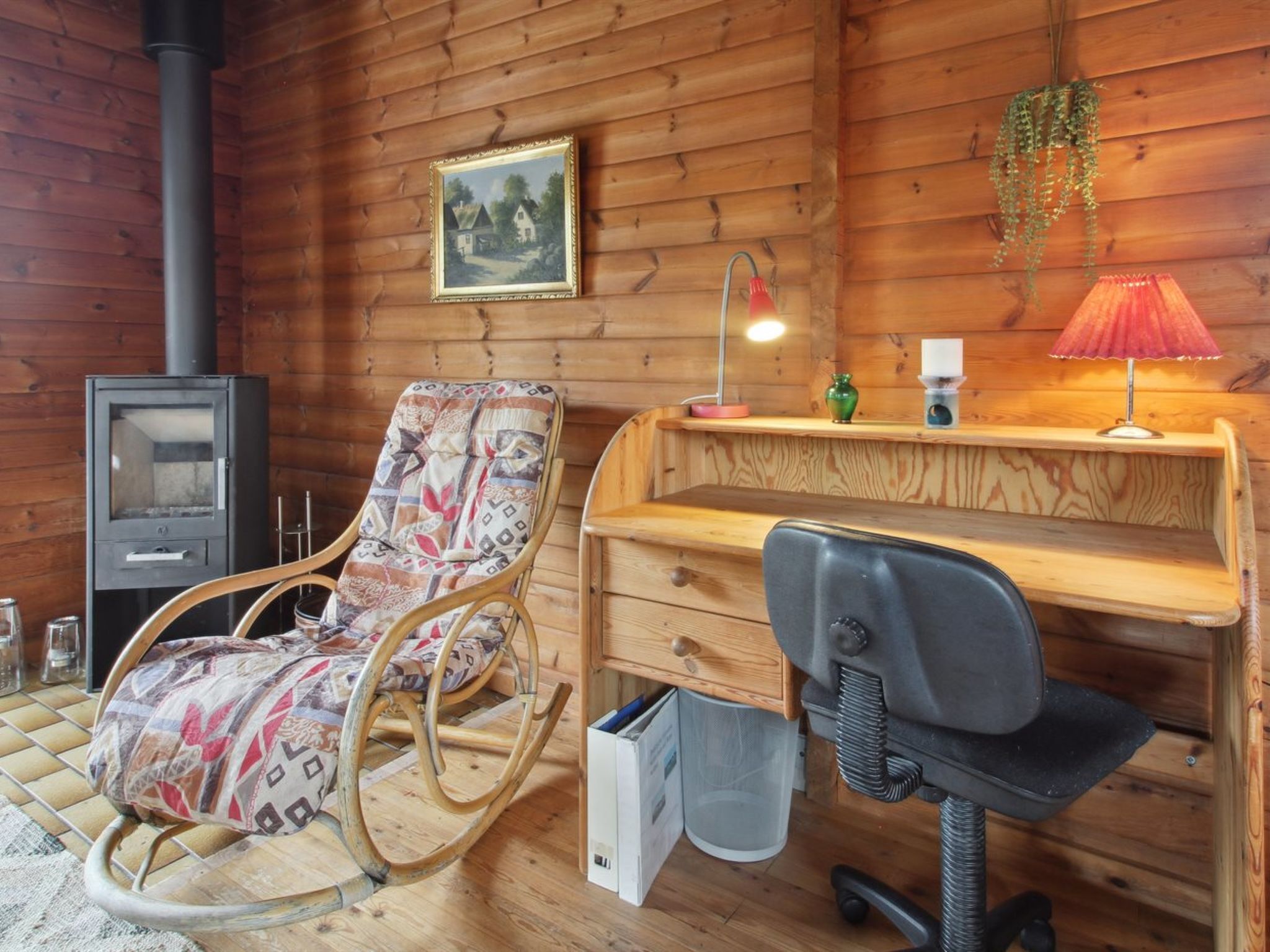 Interior acogedor de una cabaña con pared de madera, una mecedora, un escritorio con silla, un calentador pequeño y una planta colgante.